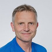 Ein lächelnder Mann mit kurzen, grauen Haaren trägt ein blaues Hemd und steht vor einem neutralen Hintergrund.