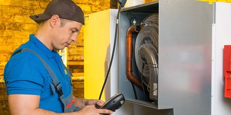 Ein Techniker in blauer Arbeitskleidung überprüft eine Maschine mit einem Handgerät in einer gut beleuchteten Umgebung.