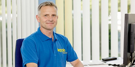 Ein Mann in einem blauen Poloshirt sitzt lächelnd an einem Schreibtisch, umgeben von hellen, vertikalen Jalousien.