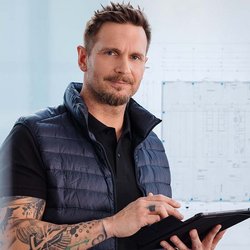 Ein Mann hält ein Tablet in der Hand und im Hintergrund ist eine Tafel an der eine Technische Zeichnung hängt