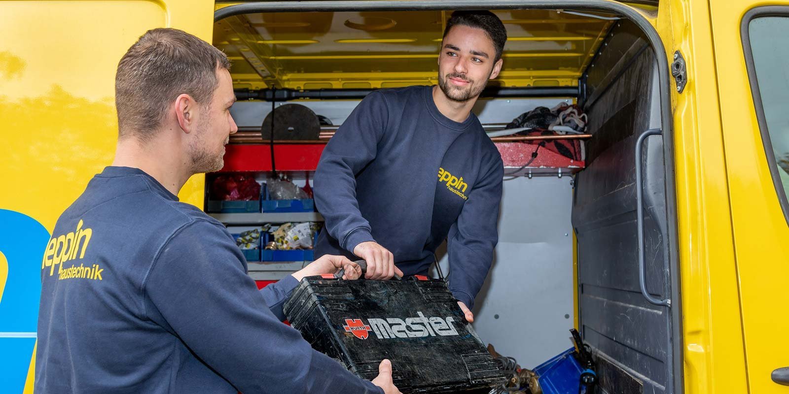 Zwei Männer in blauen Arbeitskleidung heben gemeinsam eine Werkzeugkiste aus einem gelben Lieferwagen.