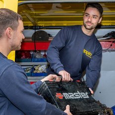 Zwei Männer in blauen Arbeitskleidung heben gemeinsam eine Werkzeugkiste aus einem gelben Lieferwagen.