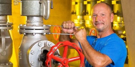 Ein Mann in einem blauen T-Shirt arbeitet an einem großen Ventil mit einem roten Handrad in einer industriellen Umgebung.