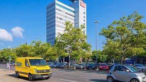 Ein gelbes Fahrzeug steht auf einem Parkplatz, während im Hintergrund ein modernes Hochhaus und blauer Himmel sichtbar sind.