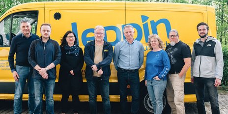 Eine Gruppe von sieben Personen steht lächelnd vor einem gelben Lieferwagen, umgeben von grünen Bäumen.