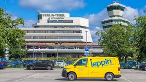 Ein gelbes Lieferfahrzeug steht auf einem Parkplatz vor dem Flughafen Berlin-Tegel, umgeben von grünen Bäumen und blauen Autos.