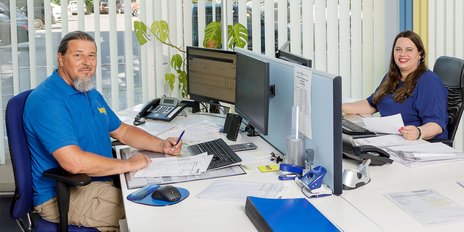 In einem hellen Büro arbeiten zwei Mitarbeiter, der Mann mit Bart trägt ein blaues Shirt, die Frau hat lange, lockige Haare.