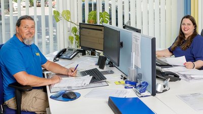 In einem hellen Büro arbeiten zwei Mitarbeiter, der Mann mit Bart trägt ein blaues Shirt, die Frau hat lange, lockige Haare.