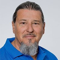 Ein Mann mit einem langen, grauen Bart und einem freundlichen Lächeln trägt ein blaues Hemd vor einem neutralen Hintergrund.