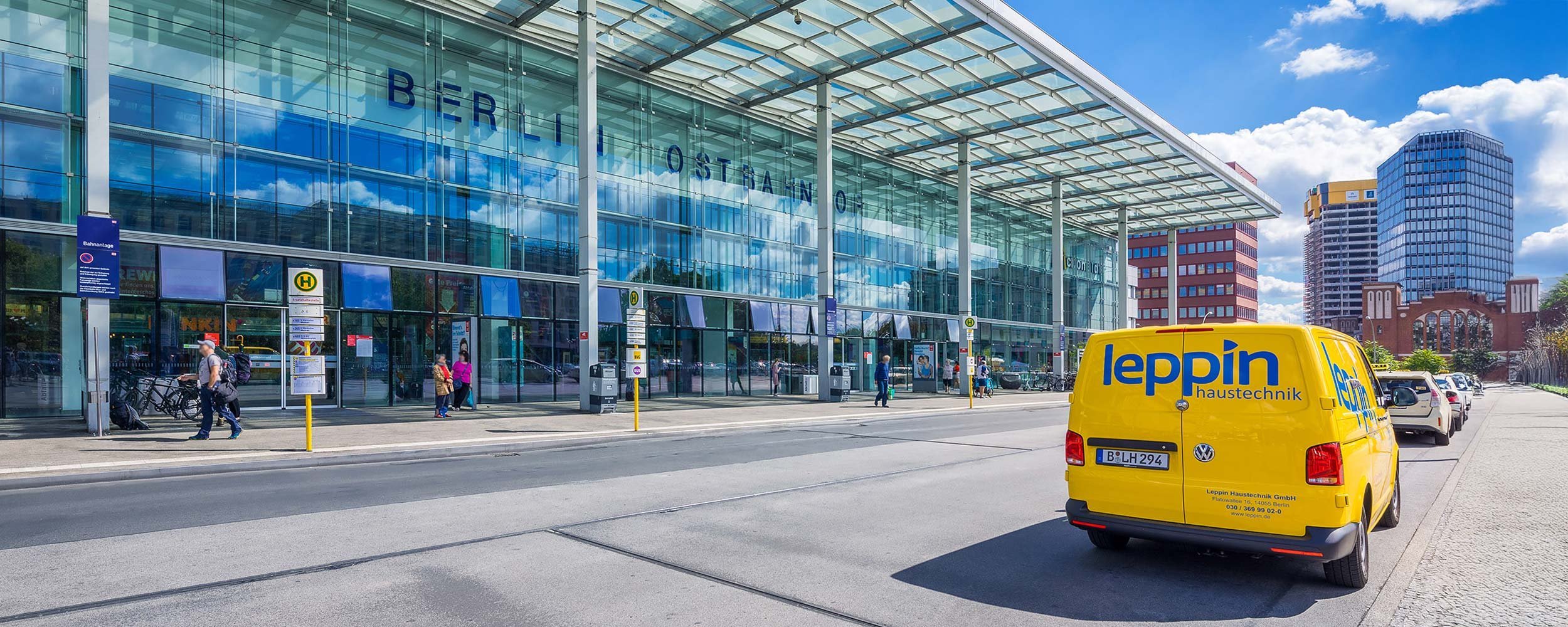 Ein gelbes Lieferfahrzeug parkt vor dem Berliner Ostbahnhof, umgeben von Menschen und der beeindruckenden Glasarchitektur.