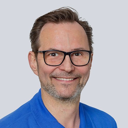 Der Mann mit Brille hat einen freundlichen Ausdruck und trägt ein blaues Oberteil, das seine positive Ausstrahlung unterstreicht.