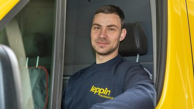 Ein junger Mann mit einem freundlichen Lächeln sitzt in einem gelben Lieferwagen und trägt ein dunkelblaues Sweatshirt.