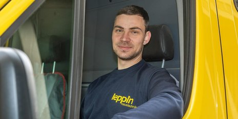 Ein junger Mann mit einem freundlichen Lächeln sitzt in einem gelben Lieferwagen und trägt ein dunkelblaues Sweatshirt.