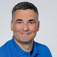 Der Mann mit einem freundlichen Lächeln hat kurzes, graues Haar und trägt ein leuchtend blaues Hemd.