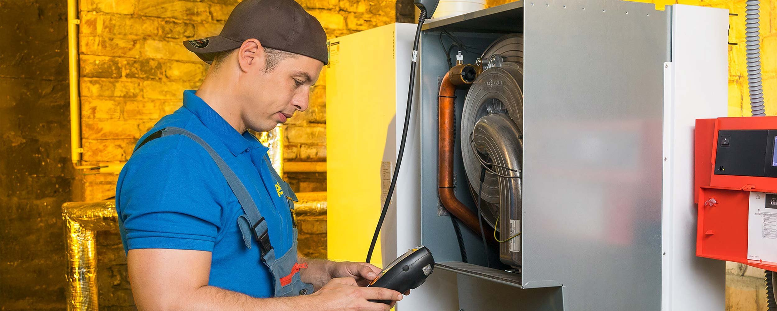 Ein Techniker in blauer Arbeitskleidung überprüft eine Heizungsanlage in einem gut beleuchteten Raum mit Ziegelwänden.